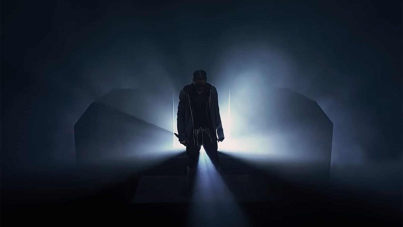 Silhouetted performer standing under a spotlight in thick fog, flanked by two dark arched stage structures.