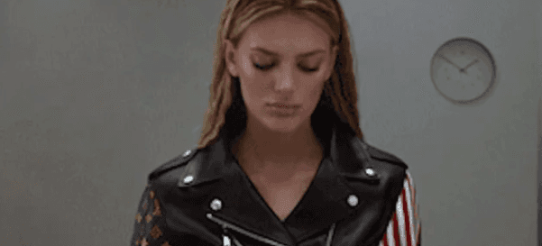 Woman in a black leather jacket standing in a room with a wall clock, looking down.