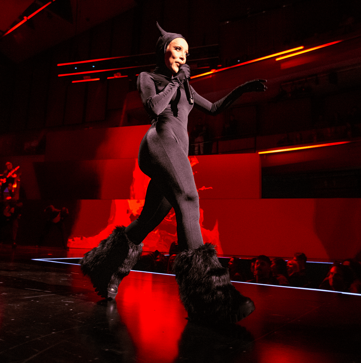 Performer in a dark catsuit with a pointed hood and furry boots striding across a red-lit stage.