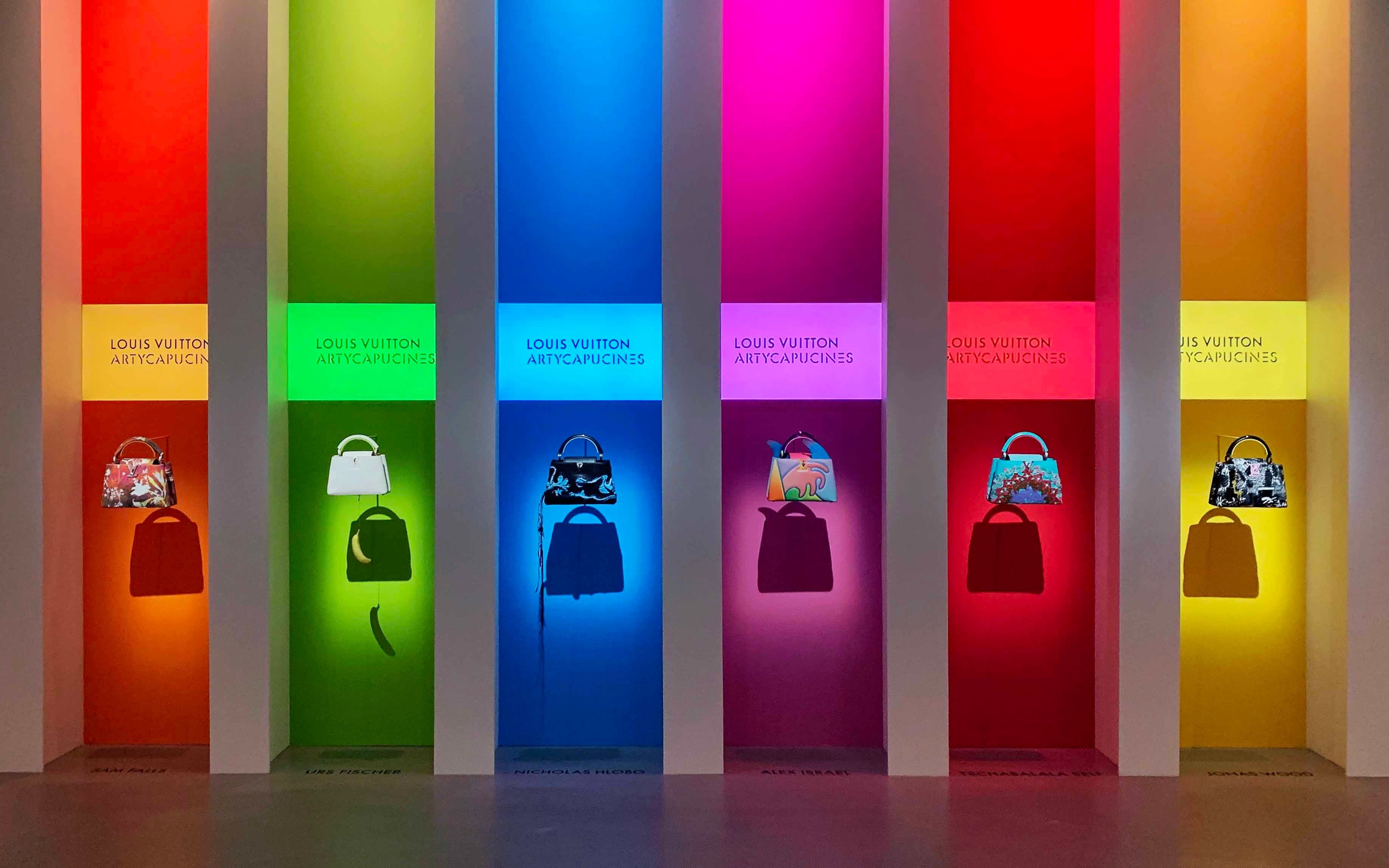 Row of tall backlit display columns in rainbow colors, each holding a small bag on a pedestal.
