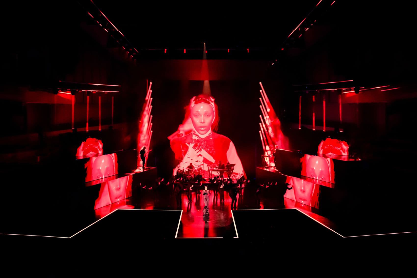 Red-lit ceremonial stage scene with a performer seated behind flowers, flanked by mirrored red screens.