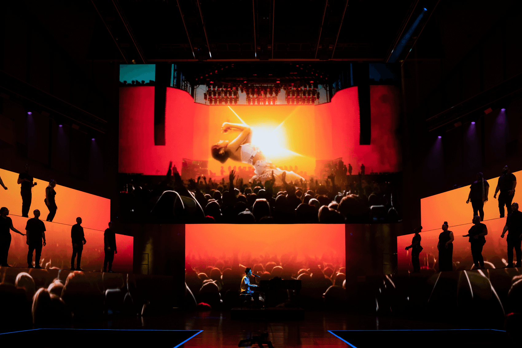 Silhouetted crowd watching a performer leap on a stage lit in orange and teal.