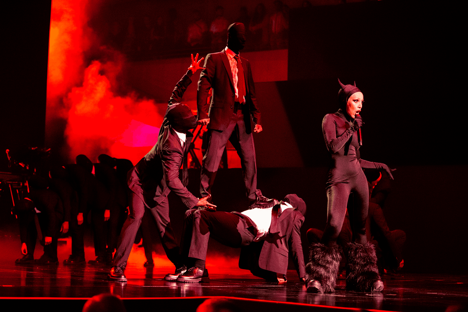 Group of dancers in dark outfits performing a coordinated pose on a red-lit stage.