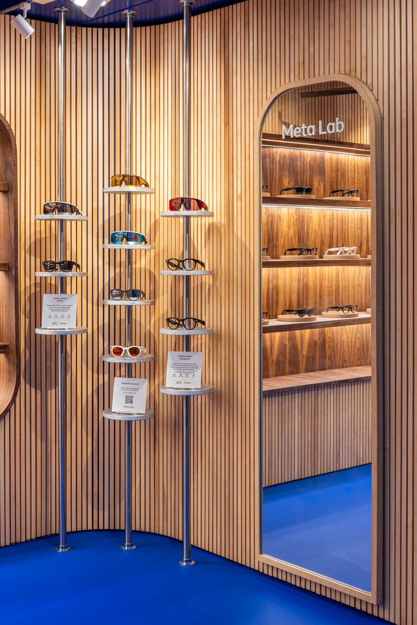 Curved wood-slat interior wall with tiered sunglasses display rods hanging in front of an arched mirror labeled 'Meta Lab'.