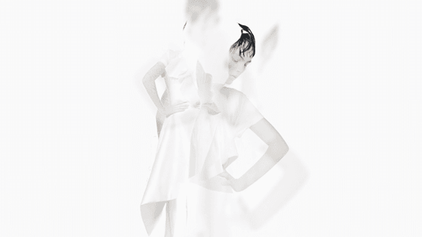 Model in a white sculptural draped dress against a white background, one hand on hip, captured in motion blur.