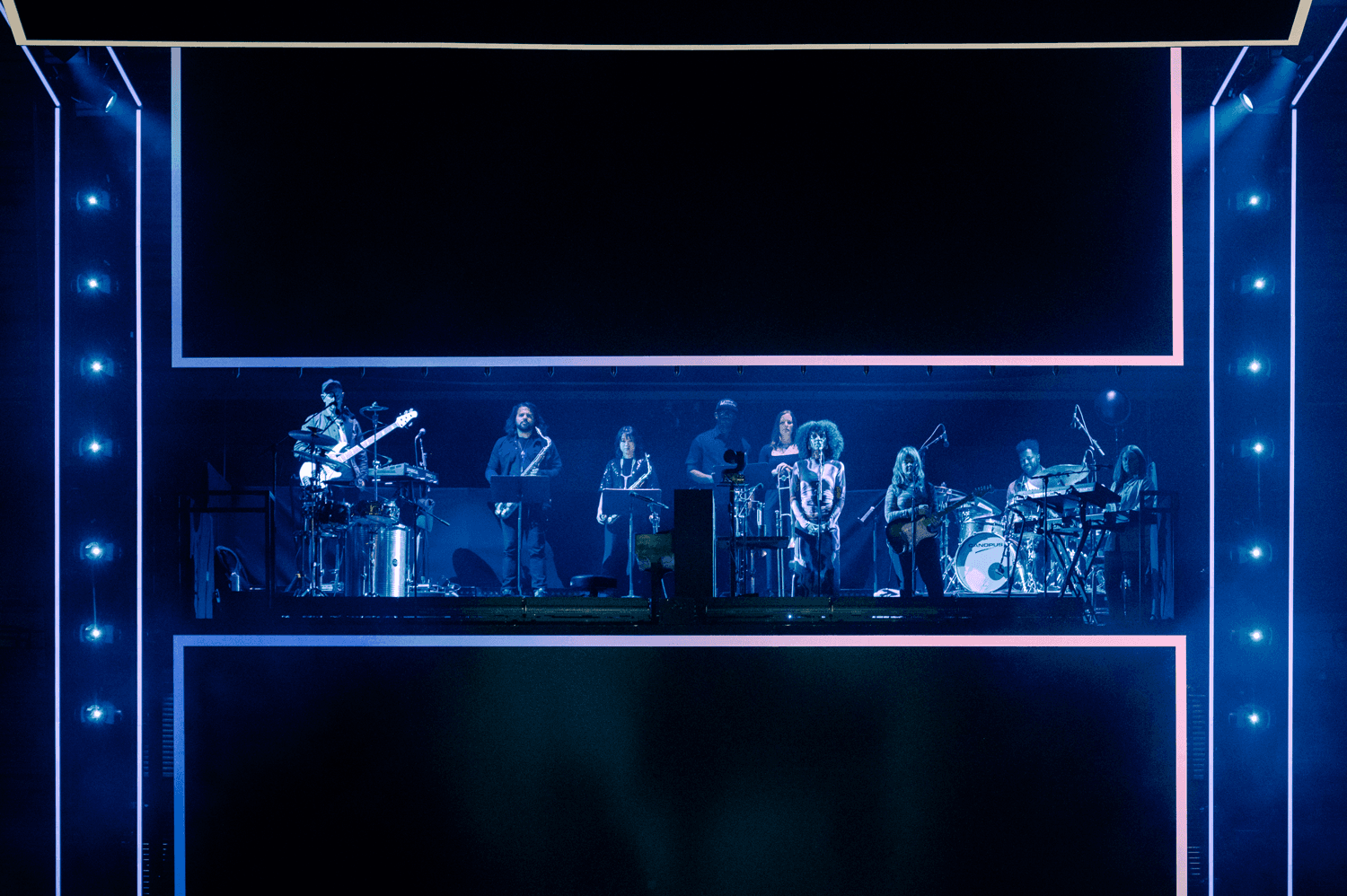 Band performing on a blue-lit stage flanked by angular screen panels.