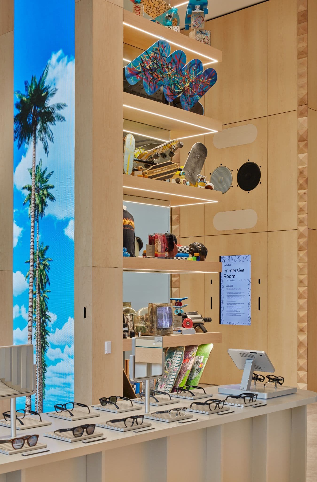 Wood display column with a backlit palm-tree graphic beside tiered shelving holding skateboards, toys, and sunglasses above a counter.
