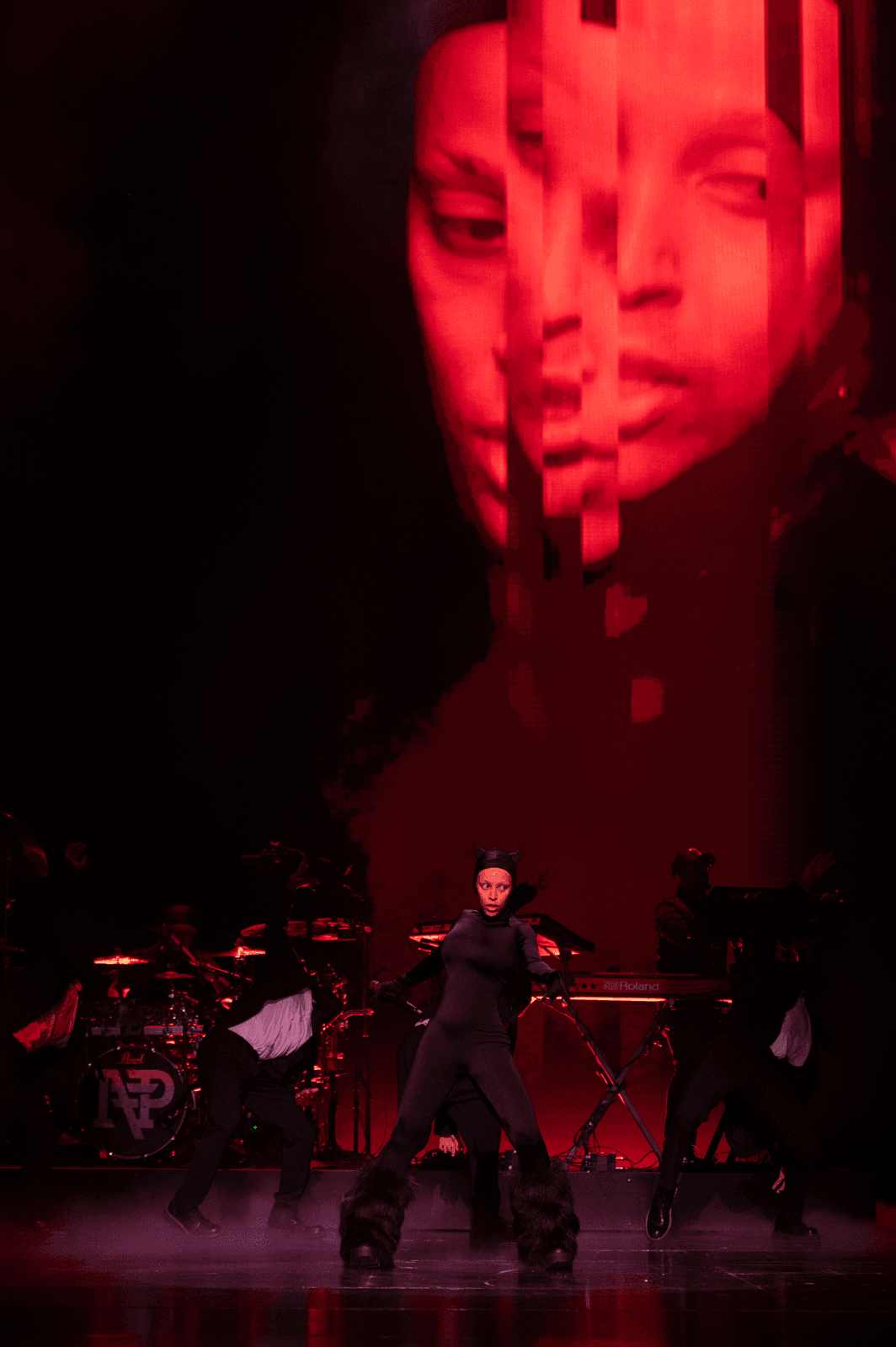Red-lit portrait with a fragmented, sliced face projection above a performer in a black catsuit performing with a band.