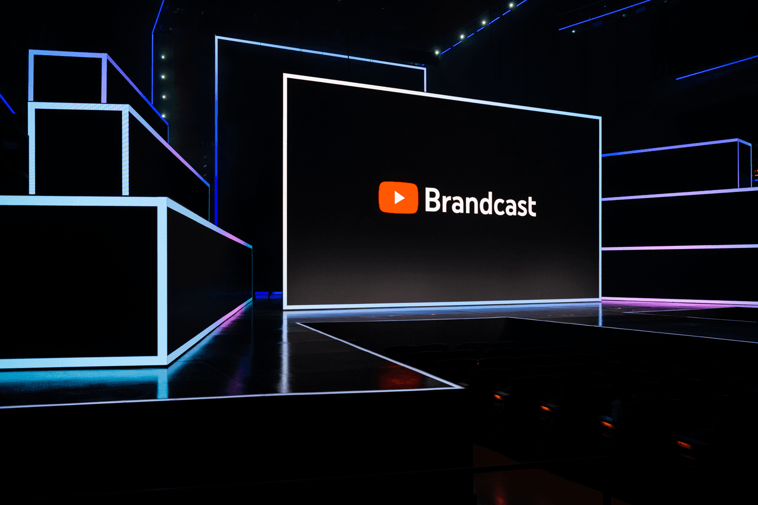 Wide shot of a blue-lit arena stage with angular LED panels and a YouTube Brandcast logo on screen.