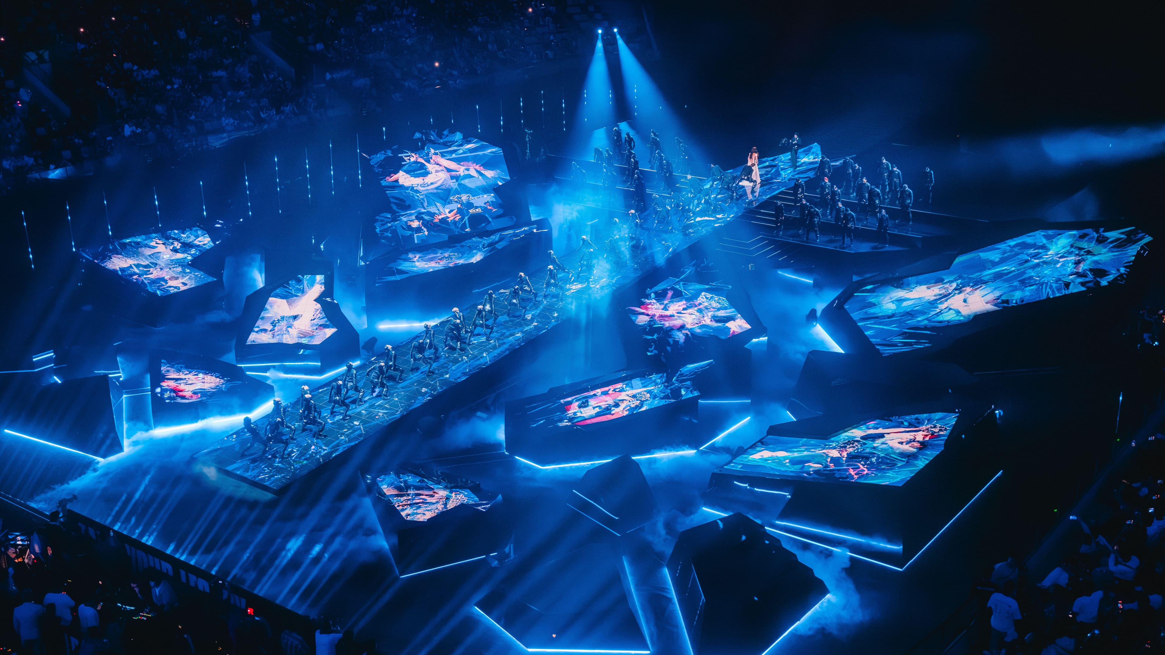 Wide stage shot with blue and red light beams converging on a circular red emblem, with the crowd visible in the arena.