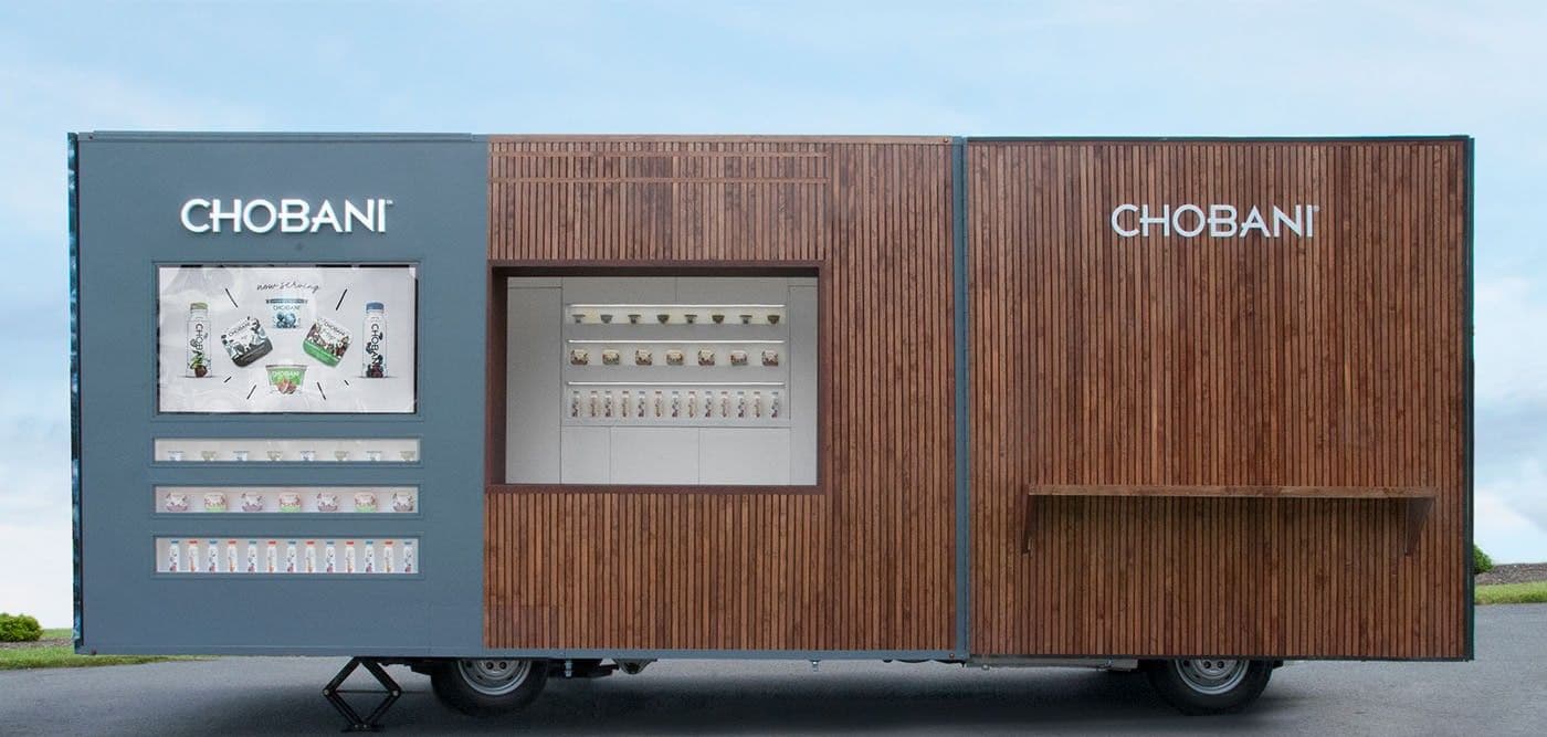 Side view of a wood-slat-clad mobile trailer branded CHOBANI, with a digital menu screen and a vending dispenser built into the facade.