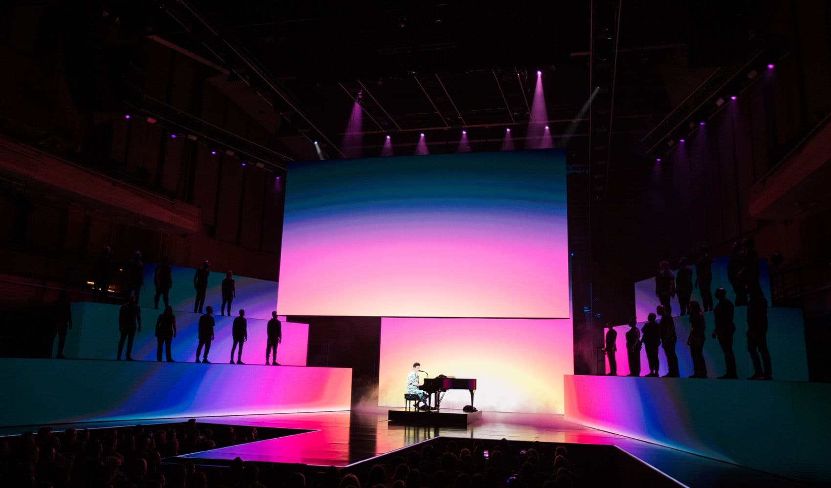 Performer at a piano on a stage lit with a pink-to-blue gradient screen.