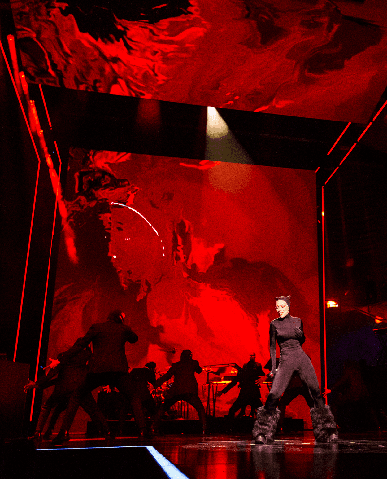 Performer in a dark catsuit posed on stage in front of a red abstract backdrop with silhouetted dancers and musicians.