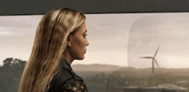 Woman in profile looking out a window at a wind turbine in a rural landscape.