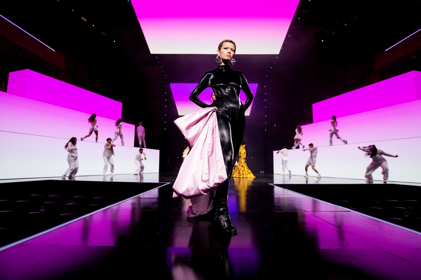 Model in a black catsuit with a pink fabric train walking a runway between pink-lit panel walls, dancers visible on side screens.