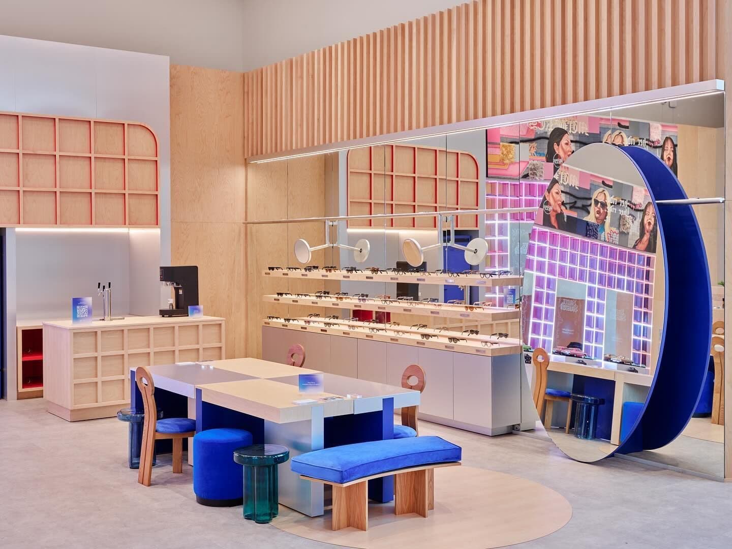 Light wood retail space with a display wall, blue geometric panel, round mirror, and a table surrounded by blue and wood stools.