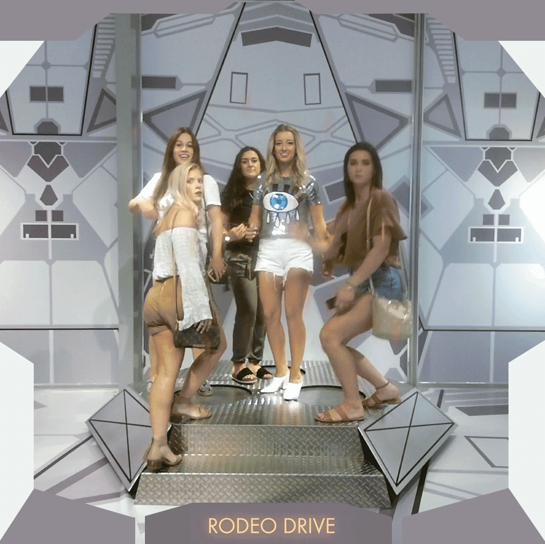 Four women posing together on a lit stage inside a geometric silver photo-booth structure, with "RODEO DRIVE" printed at the bottom.