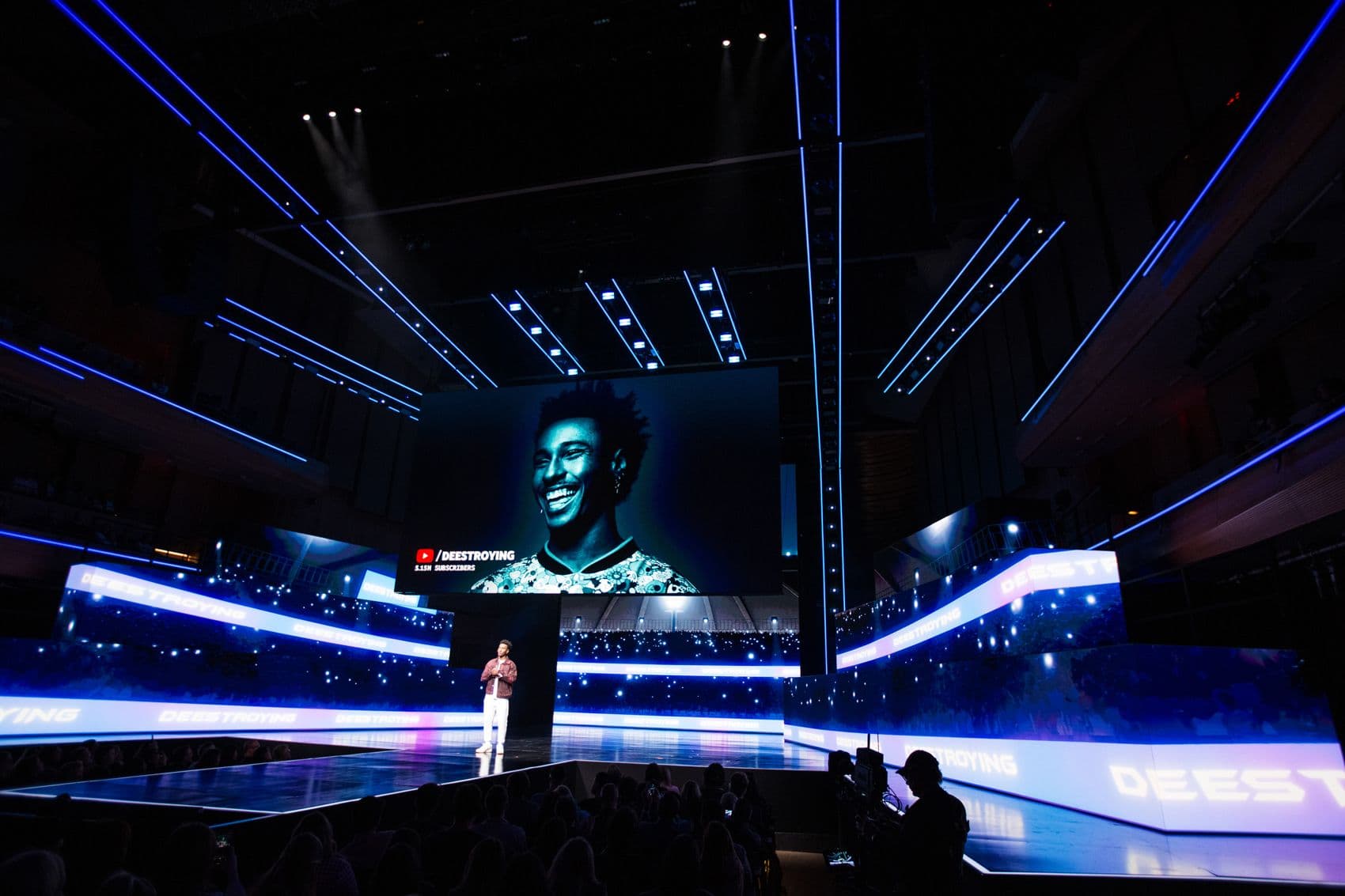 Performer on a stage with blue LED screens, seen from behind a seated audience.