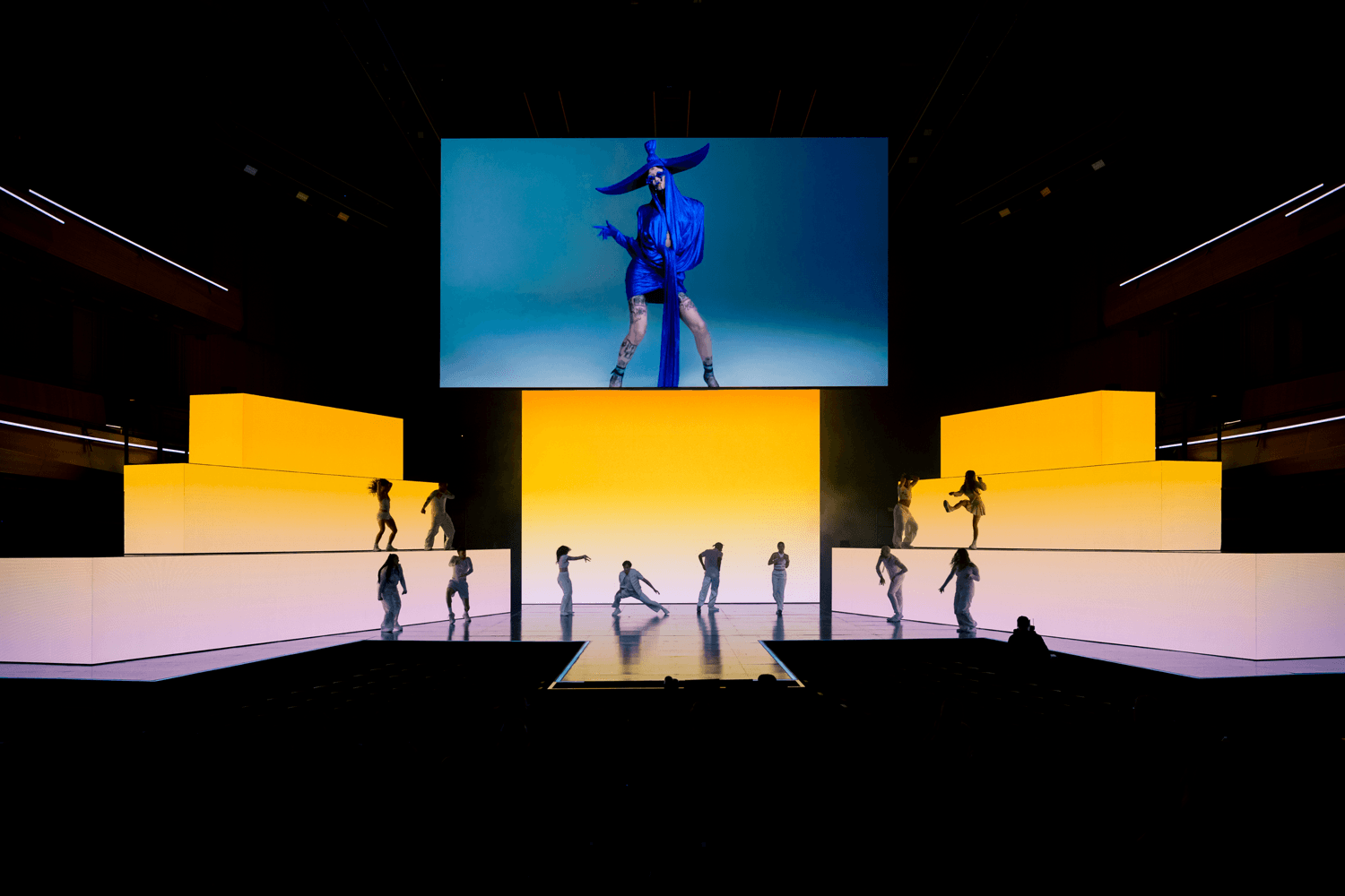 Performer in a dark outfit striding across a stage with side screens showing a dancer.